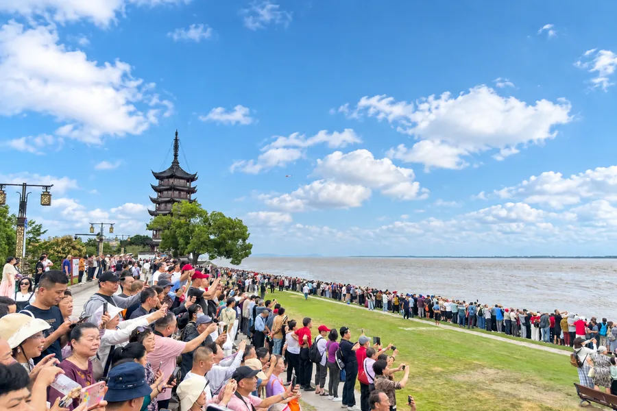 蓝天白云下，江畔古塔旁的人群沿岸观潮、举起手机记录现场